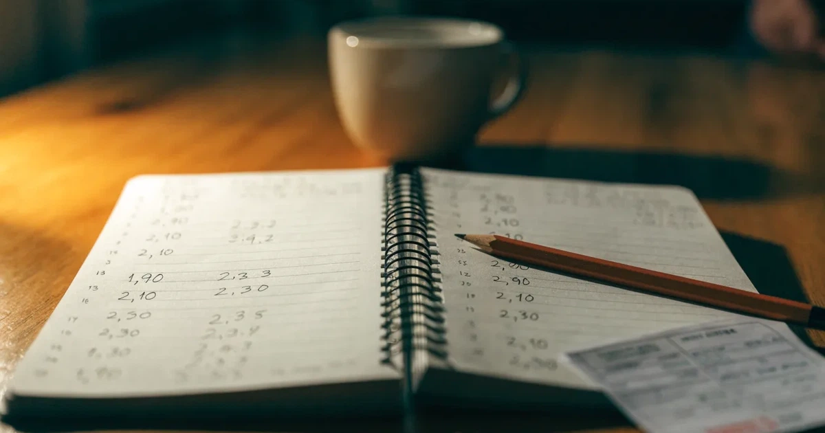 Um caderno aberto com cotas escritas à mão, uma calculadora e um lápis sobre uma mesa de madeira